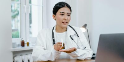 A young female doctor in a white coat on a video call, discussing medication with a patient.