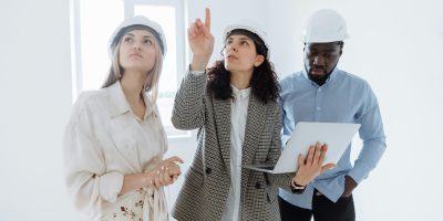 A diverse team of architects wearing helmets discusses interior plans with a laptop in a bright room.